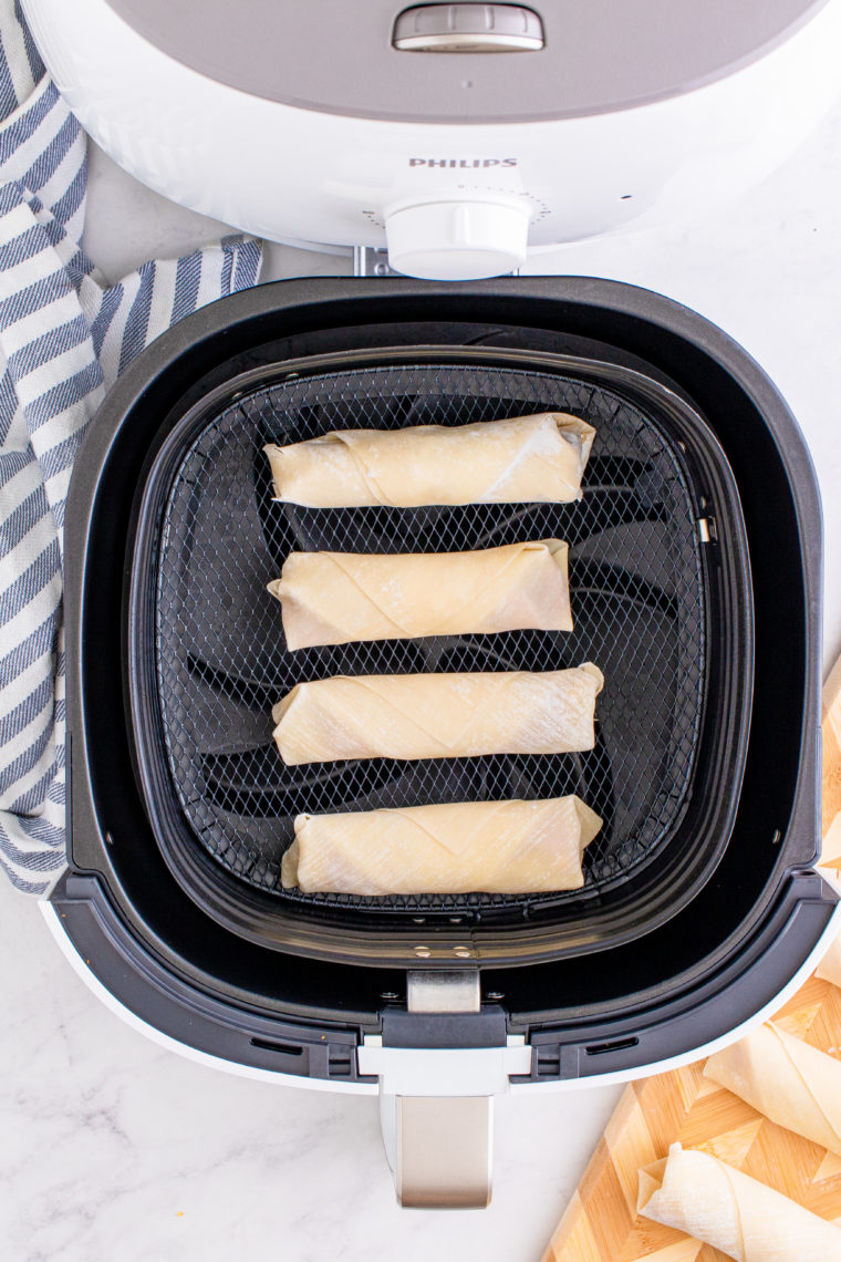 Spraying air fryer basket with nonstick spray and placing egg rolls inside, ready to cook.