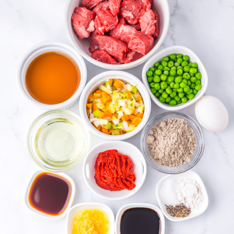 Beef stew meat, diced onion, celery, carrot, peas, tomato paste, beef broth, brown gravy mix, and a pie crust arranged on a counter