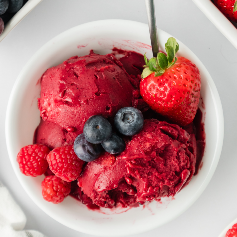  Close-up of mixed berry sorbet in a white bowl, garnished with fresh fruit and served with a spoon.