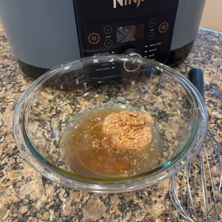 A bowl containing chicken broth, whole-grain mustard, and maple syrup being stirred together.