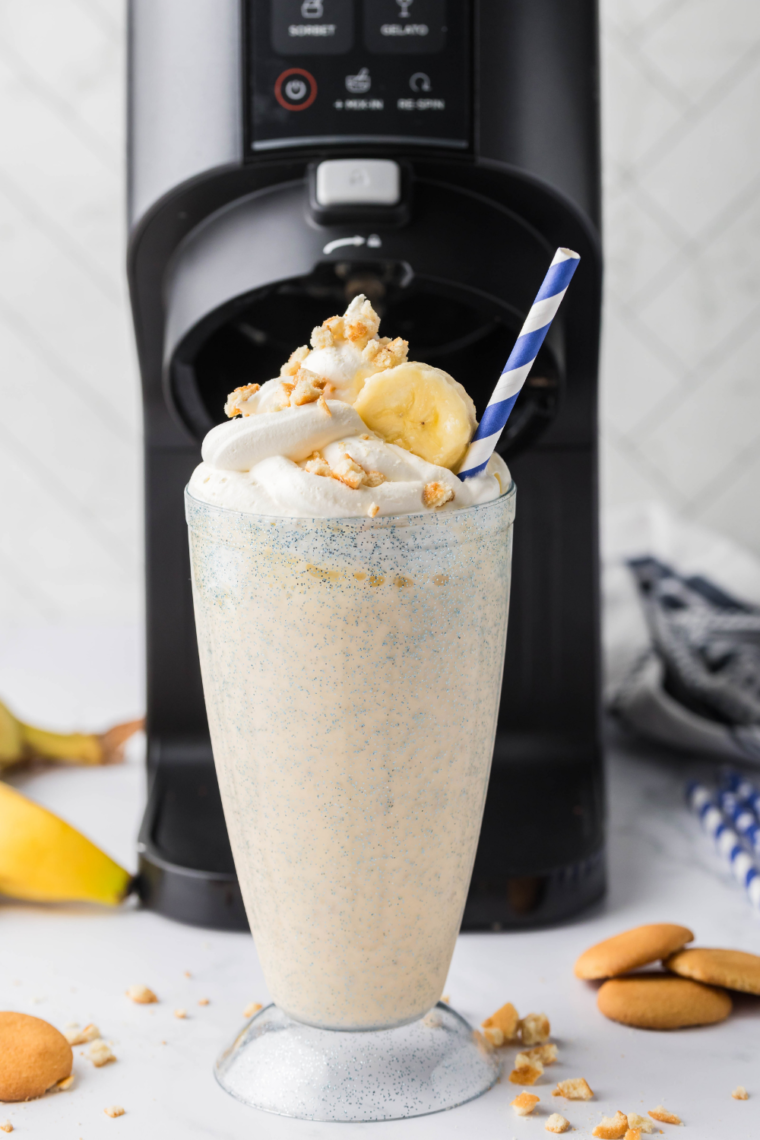 A tall glass filled with a creamy Sonic Banana Shake, topped with whipped cream and banana slices, showcasing a colorful straw alongside the Ninja Creami machine in the background.