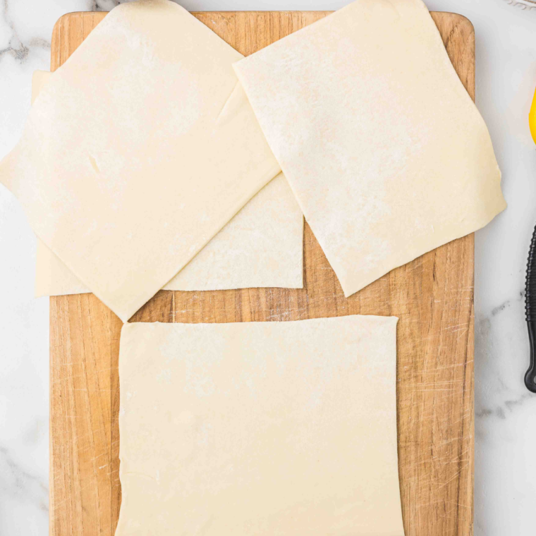 "Rolled puff pastry sheets on a floured surface, cut into four equal 6-inch squares."