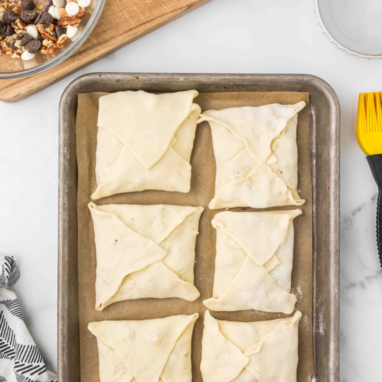 Pastry squares on a baking sheet topped with chocolate-pecan filling, edges brushed with water, and corners pinched together to seal."