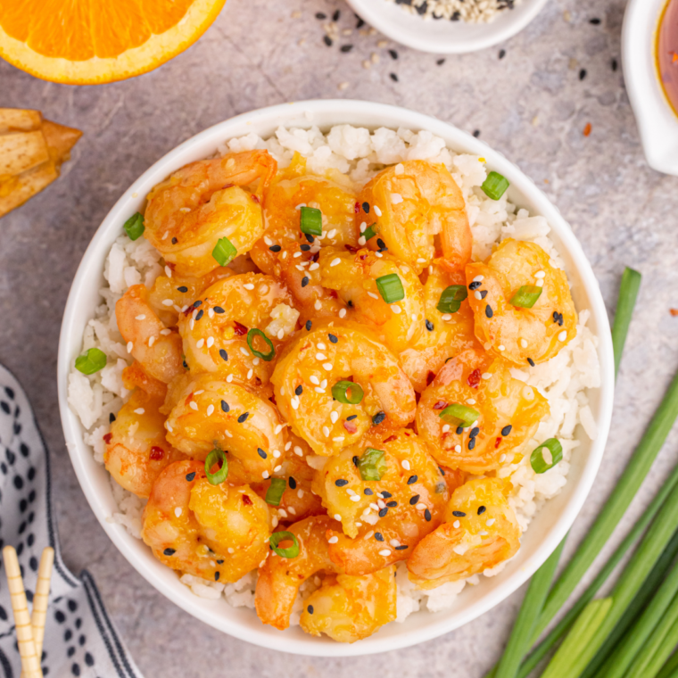 "Golden, crispy shrimp coated in a creamy, spicy dynamite sauce, garnished with sesame seeds and green onions, served on a white plate."