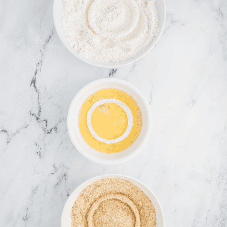 A step-by-step setup for coating onion rings: three bowls containing the flour mixture, beaten eggs with buttermilk, and panko breadcrumbs.