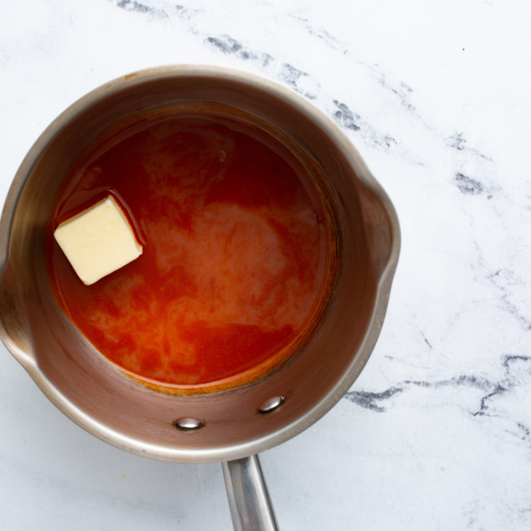 Melted butter in a saucepan with ingredients like hot sauce, cayenne pepper, garlic powder, vinegar, and paprika added, stirring together to make Wingstop Atomic Sauce.