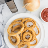 "A towering stack of crispy, golden-brown onion rings served on a plate with dipping sauces."