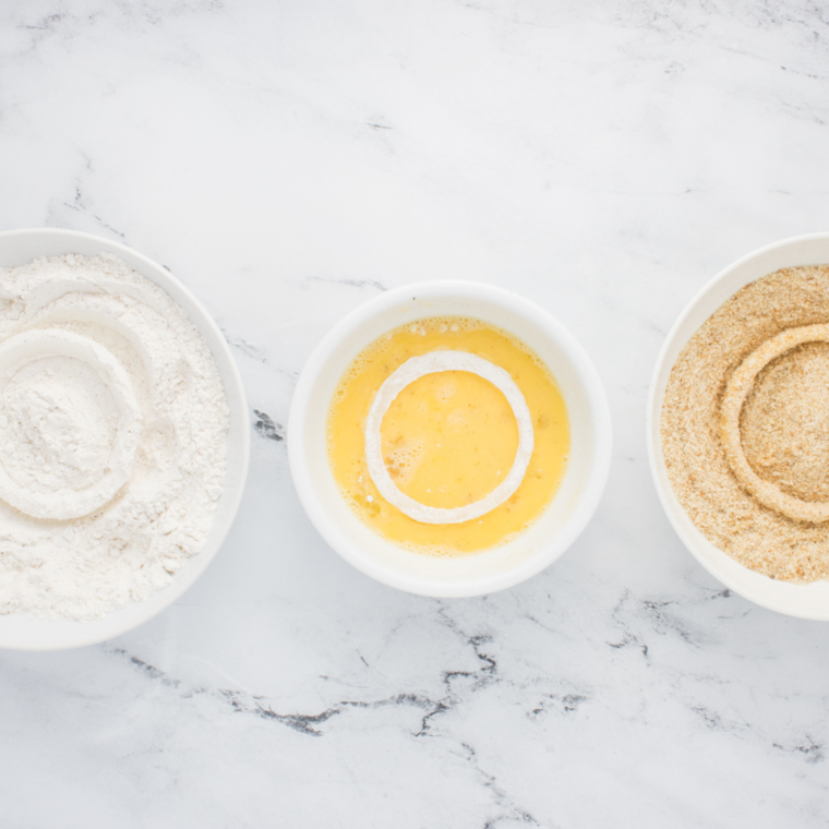 Three shallow bowls arranged with dry flour mixture, egg-milk mixture, and breadcrumbs for coating onion rings.