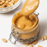 A jar of smooth, homemade peanut butter made in the air fryer, with peanuts scattered around on a wooden surface.