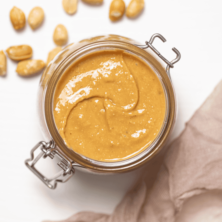 A jar of homemade peanut butter with a spoon, surrounded by roasted peanuts on a wooden surface.