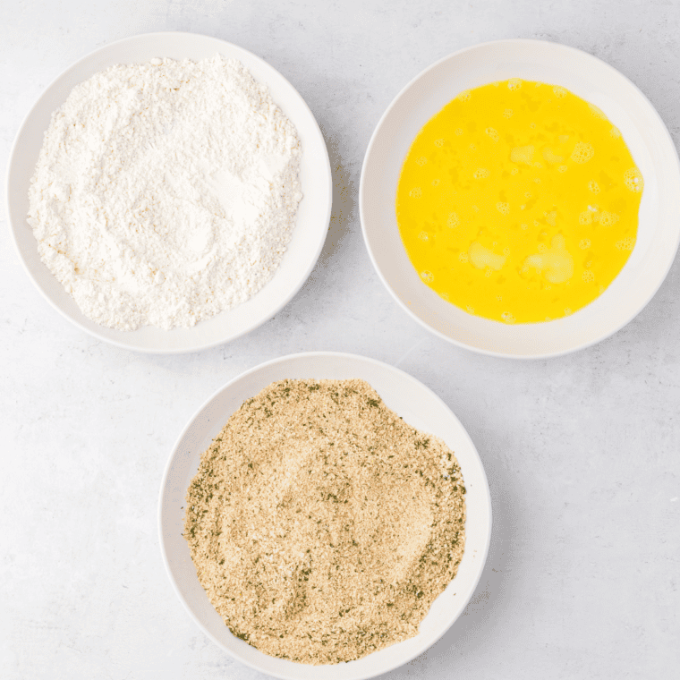Breading station setup with three shallow bowls containing flour, beaten eggs, and breadcrumb mixture for veal scaloppine.