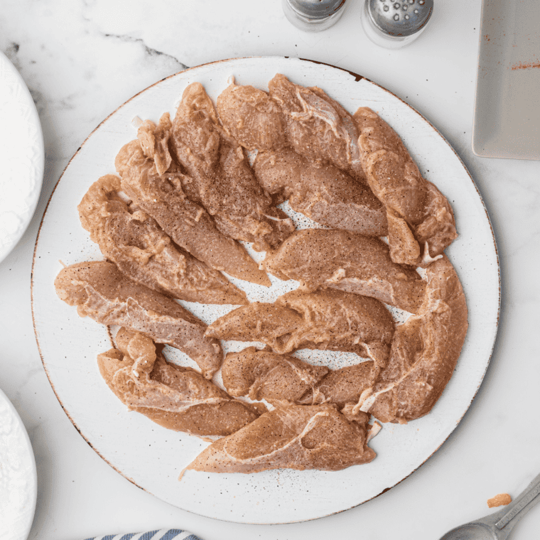 Chicken strips fully submerged in a seasoned buttermilk marinade inside a glass bowl, ready for refrigeration.