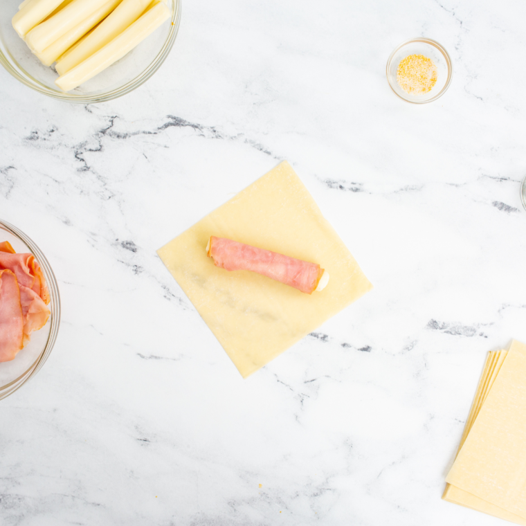 Wonton wrapper on a clean surface with a slice of ham and mozzarella stick in the center, ready to be wrapped.