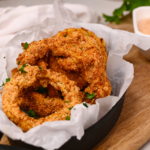 Golden, crispy keto onion rings cooked in the air fryer and served with a dipping sauce on a white plate.