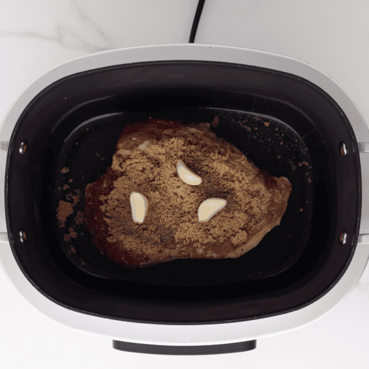 Corned beef in a slow cooker sprinkled with spices, brown sugar, black pepper, and whole garlic cloves