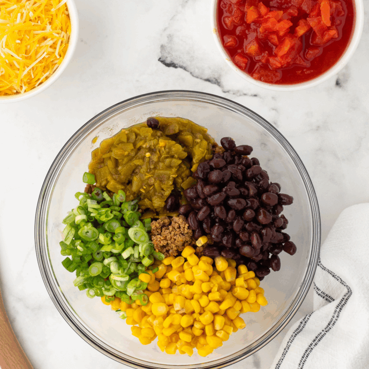 Mixing cooked ground meat, cheese, black beans, corn, tomatoes, green chilies, and green onions in a bowl, seasoning with salt and pepper.