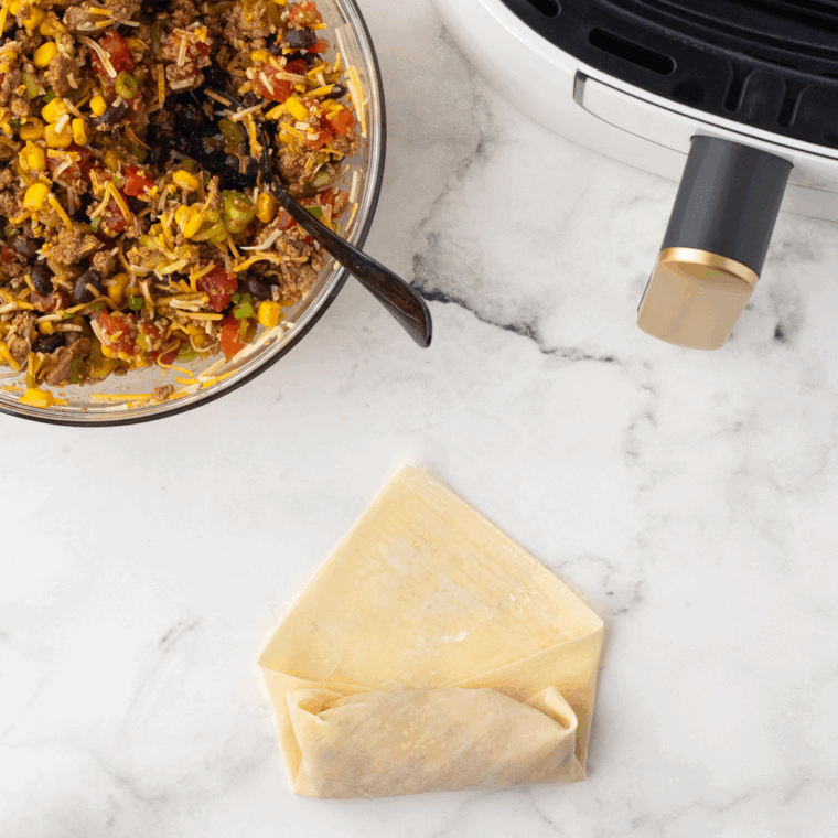 Assembling the egg rolls by placing filling on the wrapper, folding corners, and rolling tightly to seal with water.