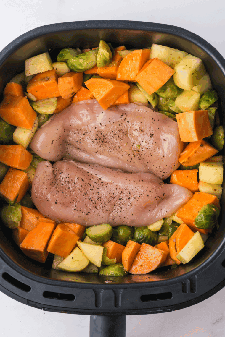 Seasoned chicken breasts placed on top of chopped vegetables in the air fryer basket.