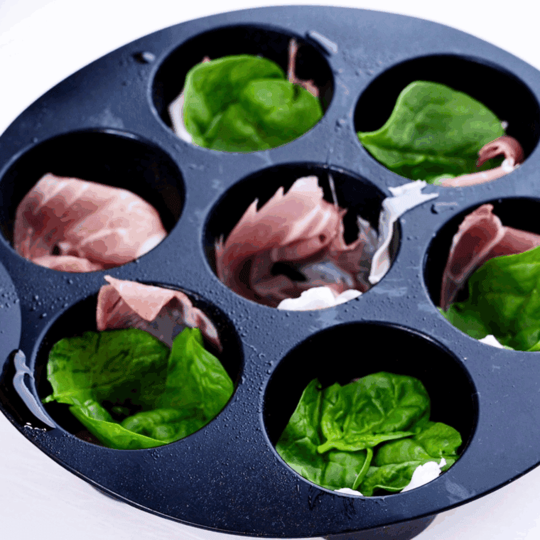 Muffin tin lined with sliced prosciutto and topped with fresh spinach leaves, ready for egg mixture.