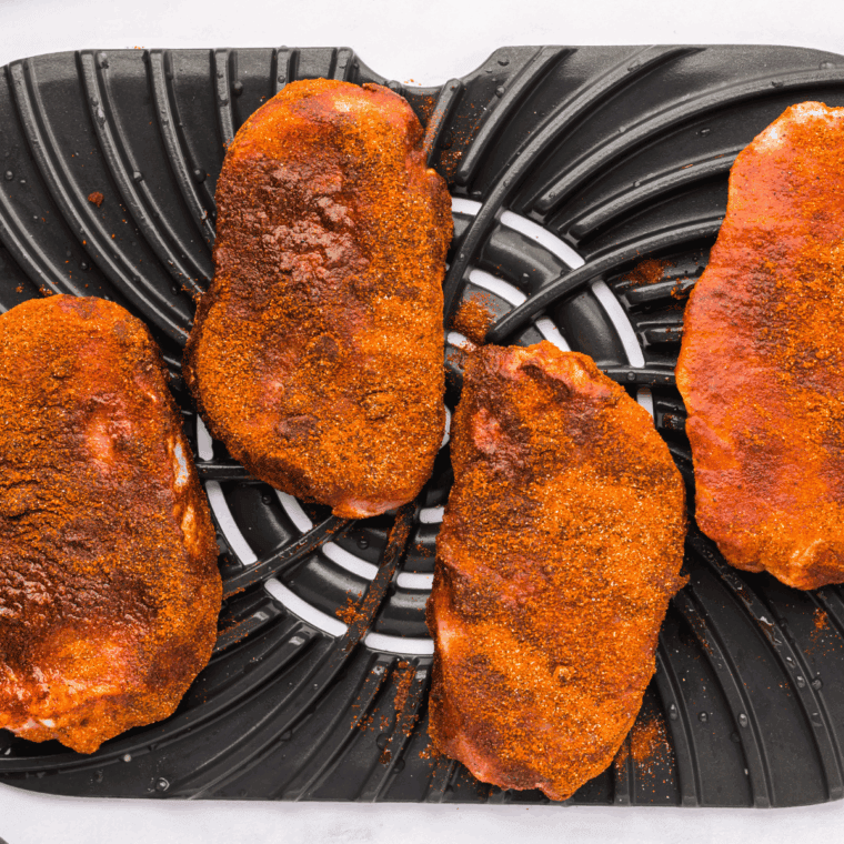 Raw pork chops being seasoned with olive oil, paprika, garlic powder, onion powder, salt, and pepper on a cutting board.