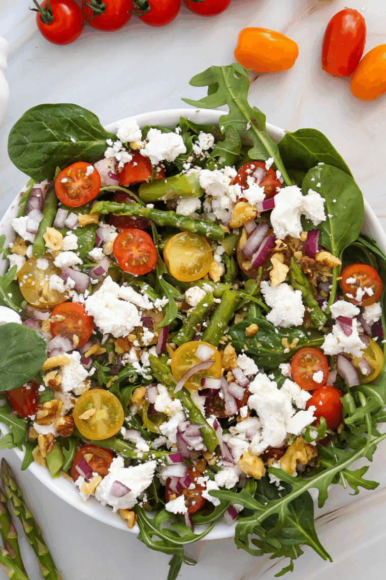 Asparagus salad with crumbled goat cheese, cherry tomatoes, red onion, and toasted nuts served in a white bowl with lemon vinaigrette.