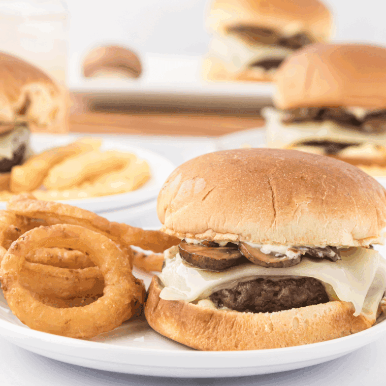 Plate of Copycat Hardee’s Mushroom Swiss Burger Recipe on kitchen table with onion rings and French fries.