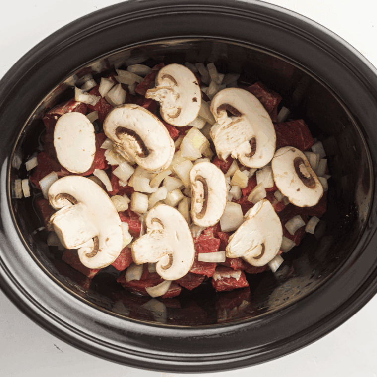 Sliced mushrooms covering beef in the slow cooker.