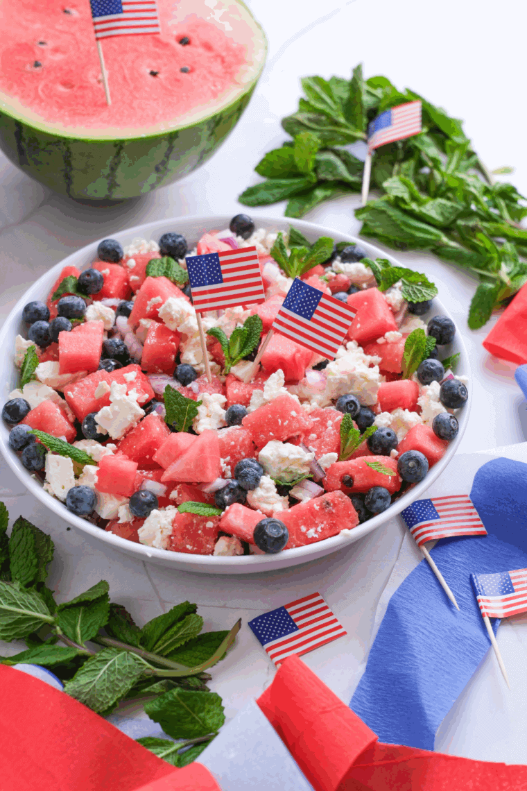 Close-up of a colorful watermelon salad with feta cheese, cucumber slices, red onion, and fresh mint leaves in a white bowl.