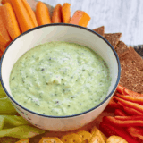 Bowl of creamy zucchini yogurt dip garnished with fresh herbs and olive oil, served with sliced vegetables for dipping.