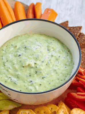 Bowl of creamy zucchini yogurt dip garnished with fresh herbs and olive oil, served with sliced vegetables for dipping.