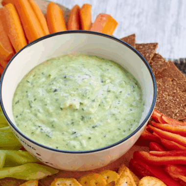 Bowl of creamy zucchini yogurt dip garnished with fresh herbs and olive oil, served with sliced vegetables for dipping.