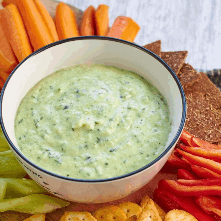 Bowl of creamy zucchini yogurt dip garnished with fresh herbs and olive oil, served with sliced vegetables for dipping.