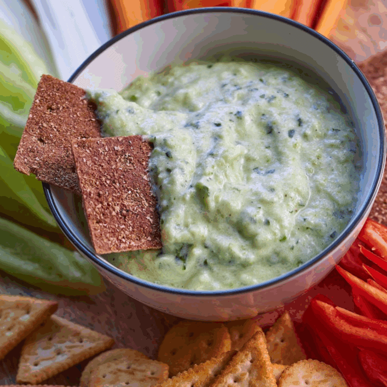 Bowl of creamy zucchini yogurt dip garnished with fresh herbs and a drizzle of olive oil, served with fresh vegetable sticks.