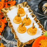 Mini pumpkin-shaped cheese balls are spread over chopped cookies, decorated with pretzel stems and parsley leaves, and placed on a fall appetizer plate.