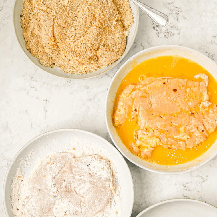 Coating chicken breast by dredging in flour, dipping in egg, and covering with breadcrumb-Parmesan mixture.
