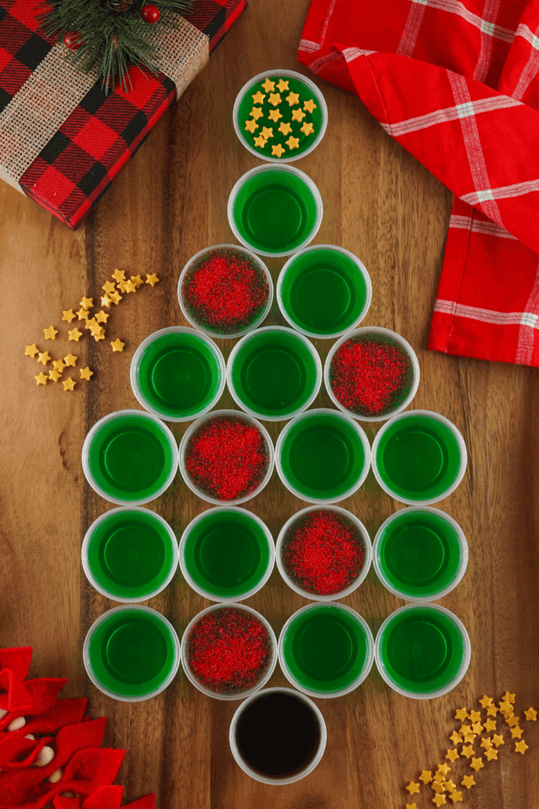 Sparkling Christmas tree Jello shots with green layers, gold star sprinkles, and red sugar ornaments arranged on a festive holiday tray.