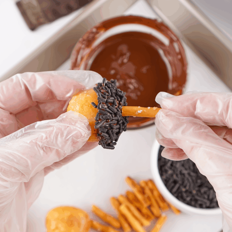 Gently pressing a pretzel stick into the chocolate-topped donut hole to form acorn stem.