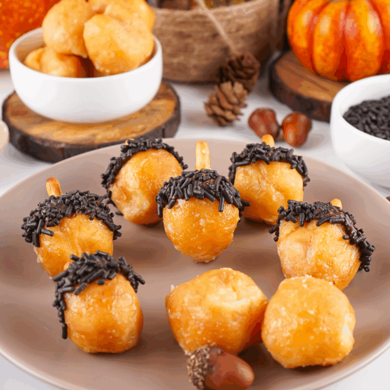 Plate of bite-sized acorn-shaped donut holes decorated with chocolate, sprinkles, and pretzel stems.