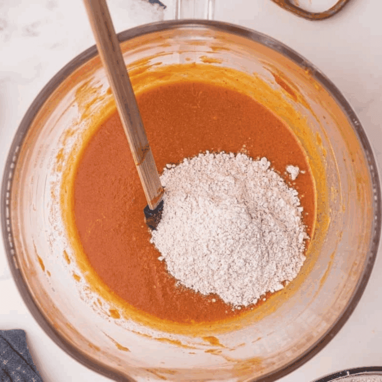 Whisking pumpkin purée, sugars, oil, eggs, milk, and vanilla extract in a bowl to create a smooth and creamy batter for pumpkin bread.