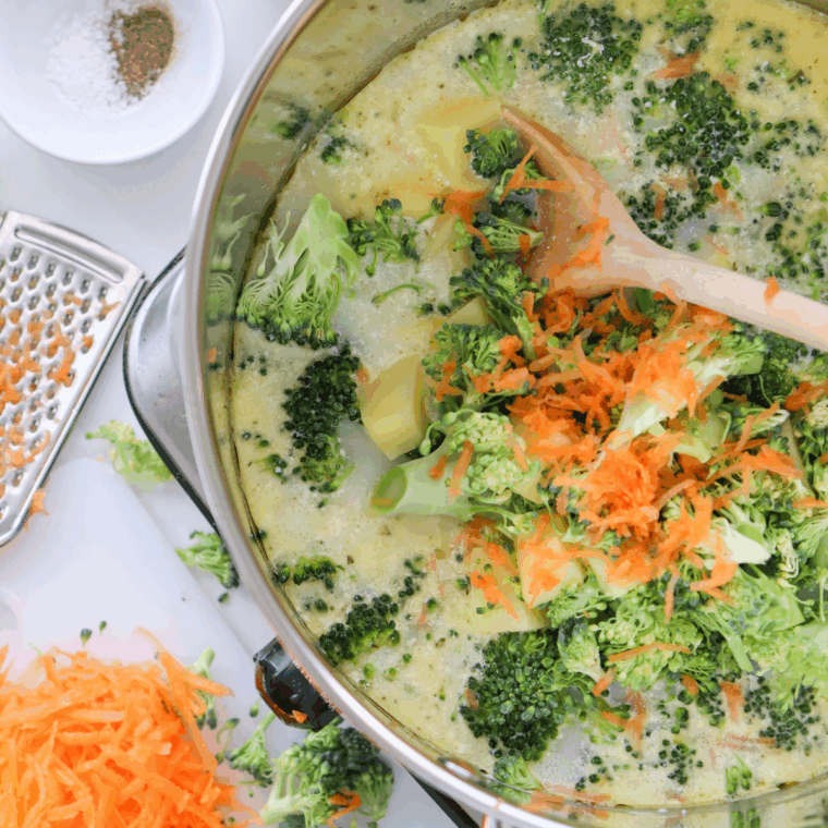 Fresh broccoli florets and shredded carrots simmering in a creamy mixture inside a soup pot.