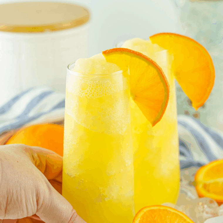 Glass of frozen mimosa made with orange juice and sparkling wine, garnished with an orange slice and served with a straw.