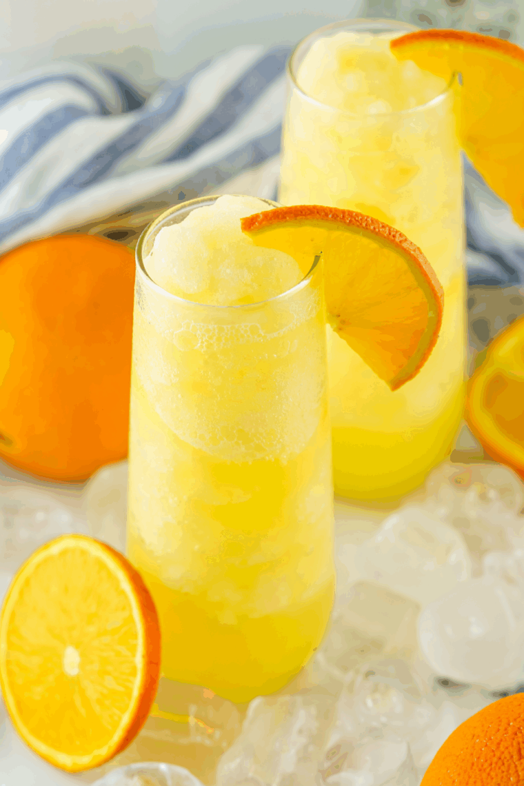 Frosty glass of frozen mimosa made with orange juice and champagne, garnished with an orange slice and bubbly foam.