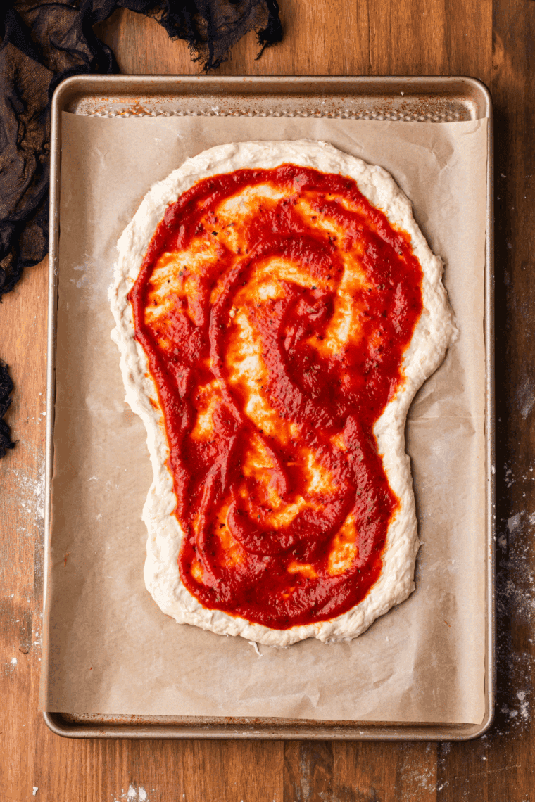 Spreading pizza sauce evenly over the skull-shaped dough, leaving a crust border.