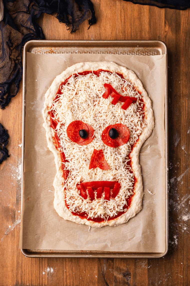 Sprinkling shredded mozzarella over sauce and arranging pepperoni, olives for eyes, red pepper strips for mouth.