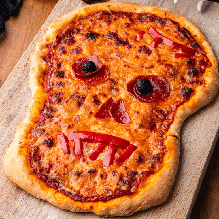 Skull-shaped Halloween pizza decorated with cheese and pepperoni on a baking sheet.