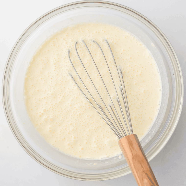 Large mixing bowl with heavy cream, milk, sugar, vanilla, lemon juice, and salt being whisked, with macerated strawberries stirred in.