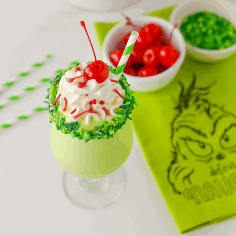 Festive green Grinch milkshake in a glass, topped with whipped cream, green and red sprinkles, and a maraschino cherry.