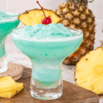 Glass of Blue Hawaiian Cocktail topped with pineapple slice and cherry, served over ice with a tropical background.