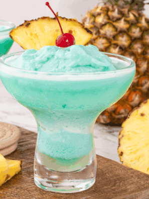 Glass of Blue Hawaiian Cocktail topped with pineapple slice and cherry, served over ice with a tropical background.