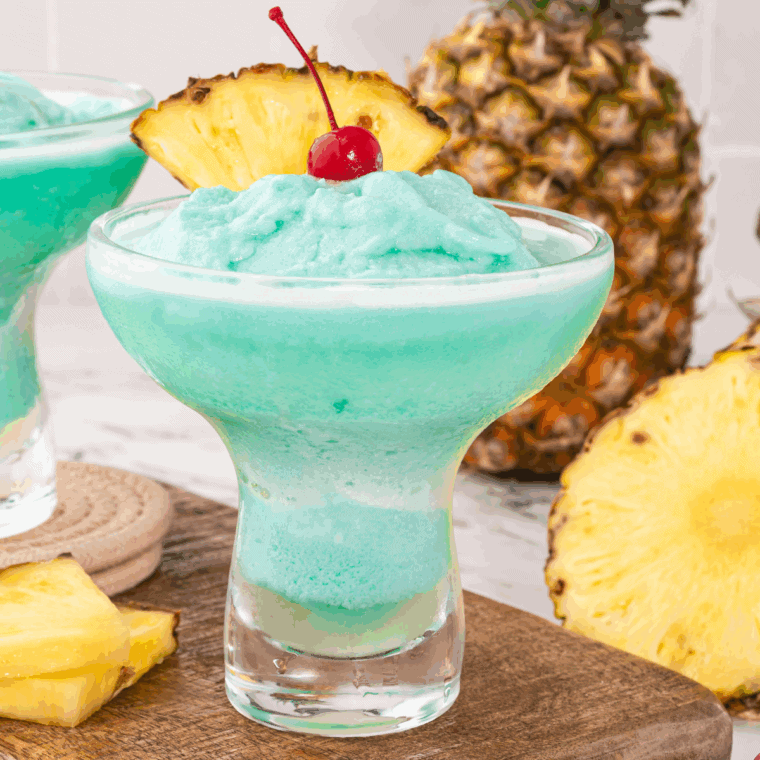 Glass of Blue Hawaiian Cocktail topped with pineapple slice and cherry, served over ice with a tropical background.